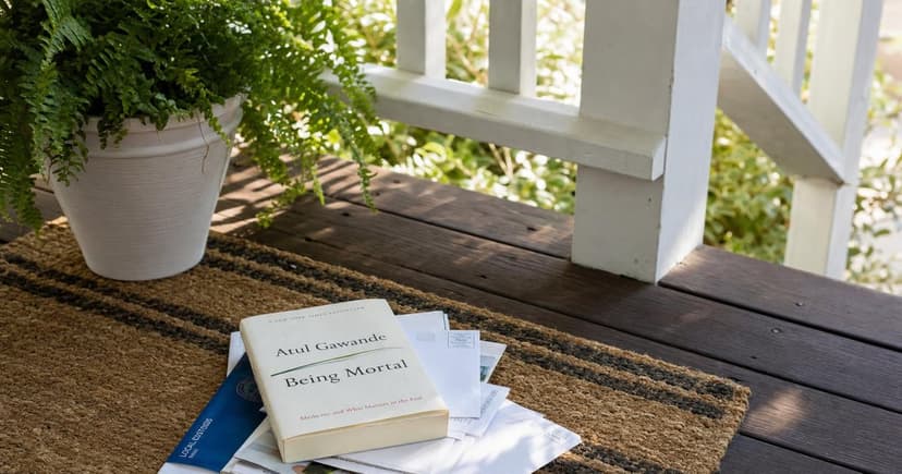 A wooden front porch step adorned with a cozy, woven rug, a potted fern in the corner catches the afternoon light, casting soft shadows, while a well-used paperback rests atop a stack of mail waiting to be sorted.